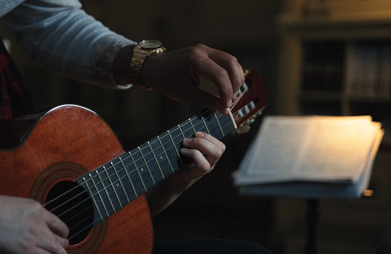 A tutor's hands adjusting a music stand beside an acoustic guitar in a lamp-lit teaching room