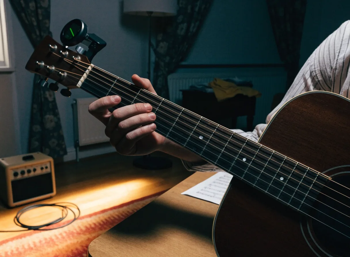 A close crop of fingers fretting a chord, with a tuner on the headstock and a practice amp behind