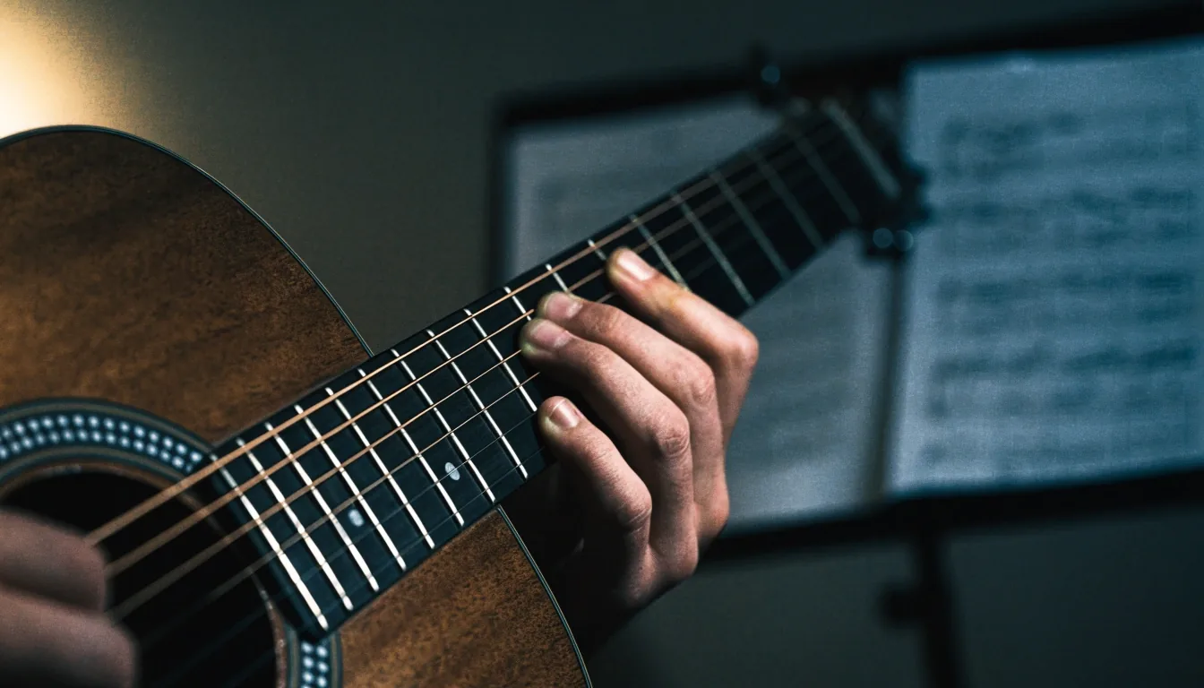 Close crop of a learner's fingers pressing open chords on an acoustic guitar in a dim practice room